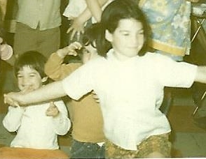 A little girl sitting on the floor laughing, a partially obscured little boy behind her making a monster face and a bigger girl with her arms out in a "ta-da" gesture.